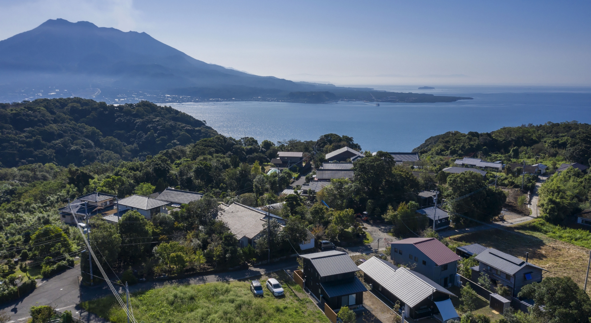 空から見下ろした穏やかな青い海と緑の山、木々に囲まれた集落が広がる沿岸の風景の様子。