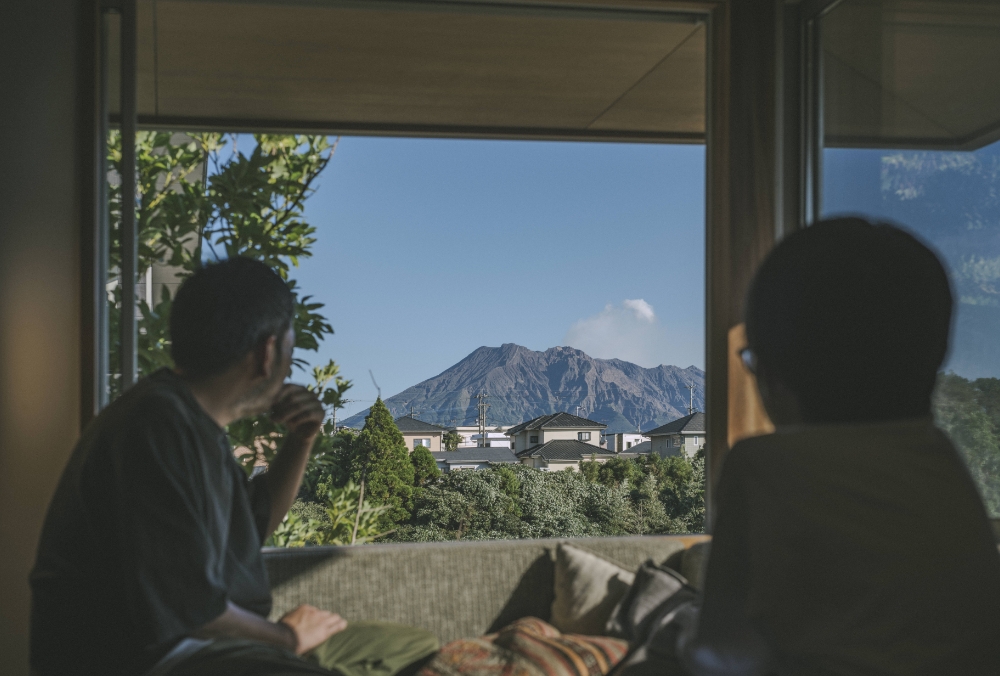 窓越しに望む雄大な山々と、緑に囲まれた住宅を眺めながらお茶をする夫婦の様子。
