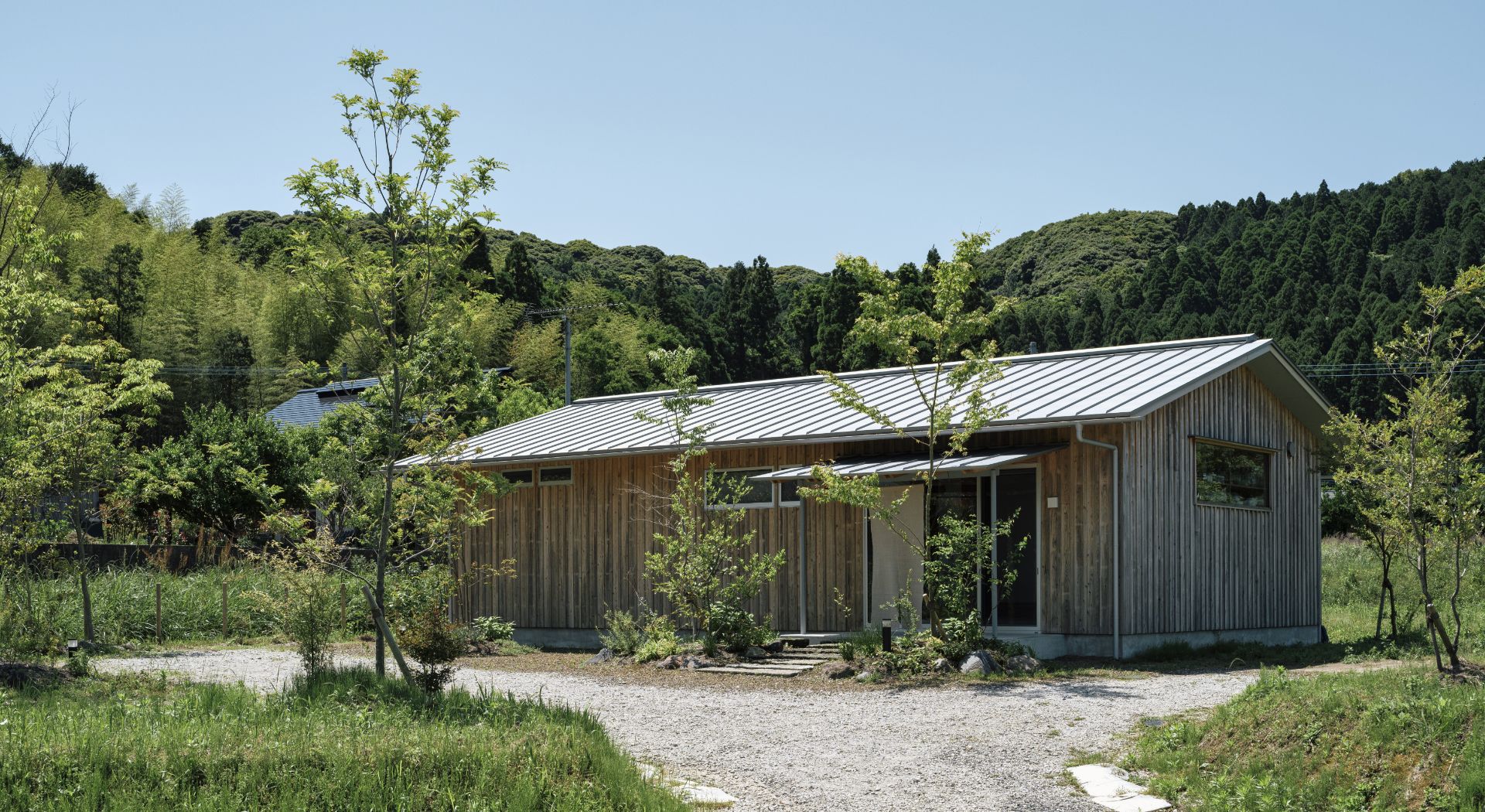 青空と緑豊かな山々に囲まれた木目調のモダンな平屋。砂利道が玄関へ続く穏やかな風景の様子。