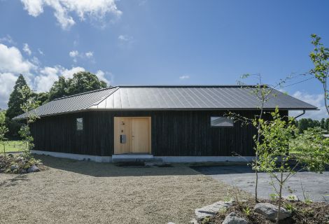 角張った形の黒い屋根のモダンな平屋。砂利の庭と緑、青空に映える外観の様子。