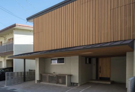 上階を薄茶色の木製ルーバー、下階をグレー外壁が覆う二階建て。車庫と木製ドアの玄関が映る外観の様子。