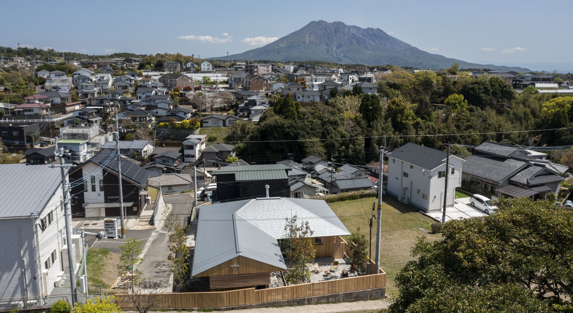 上空から見下ろした住宅街の眺望。手前に木製のモダンな平屋、奥に雄大な桜島を望める光景