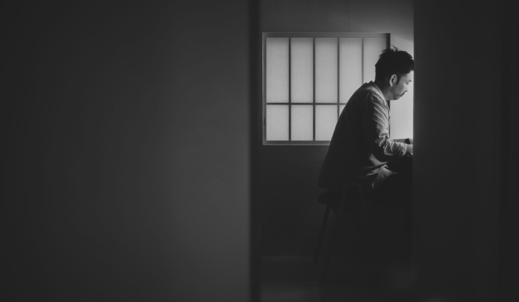 格子窓から柔らかな光が差し込む部屋。窓際で静かに読書をする男性の横顔が捉えられた白黒写真